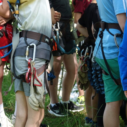 Kinder stehen mit Klettergurten und Sicherheitsausrüstung, darunter Karabiner und Handschuhe, in einer Gruppe zusammen. Im Hintergrund sind weitere Teilnehmer und Bäume zu sehen.