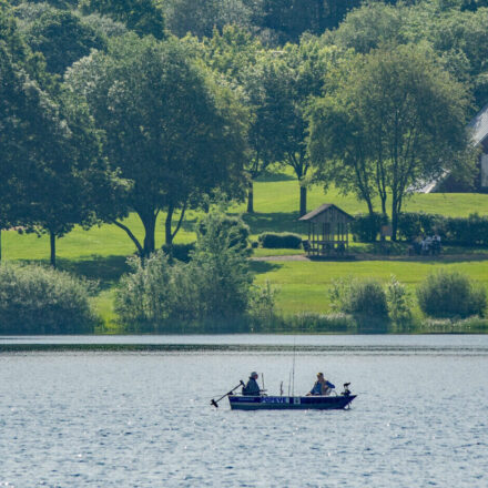 Idyllic scene on Lake Bütgenbach: two people fishing in a small boat floating on the glistening surface of the water. In the background is a green landscape with trees, a small building and a chalet, nestled in the peaceful natural surroundings. The forest in the background rounds off the harmonious scenery.