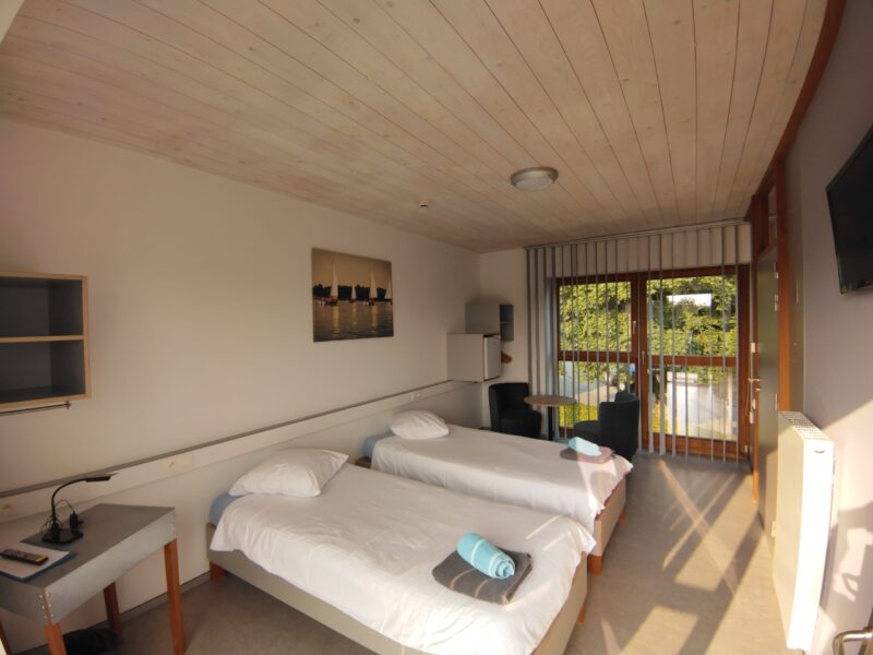 Chambre avec des lits dressés, deux fauteuile, une petite table, une télévision, un réfrigérateur