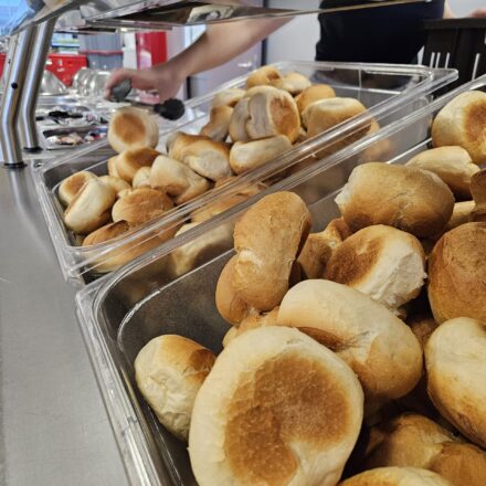 Selbstbedienungstheke mit frischen, goldbraun gebackenen Brötchen in durchsichtigen Behältern. Im Hintergrund greift eine Person mit einer Zange nach einem Brötchen.