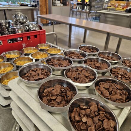 Ein Selbstbedienungsbereich einer Cafeteria mit einem Frühstücksangebot. Im Vordergrund stehen zahlreiche kleine Metallschalen, gefüllt mit Cornflakes und Schoko-Cerealien, ordentlich auf Tabletts gestapelt. Im Hintergrund ist ein großer, heller Raum mit großen Fenstern zu sehen. Dort befinden sich mehrere Getränkeautomaten, Gläserregale und ein Bereich mit Kaffeetassen und Zubehör. Die Umgebung wirkt aufgeräumt, funktional und auf eine größere Gästezahl ausgelegt.