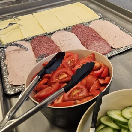 Ein Frühstücksbuffet in einer Kantine mit einer Auswahl an Aufschnitt und frischem Gemüse. Im Vordergrund liegt eine silberne Platte mit ordentlich aufgefächerten Scheiben Käse, Salami und Lyoner. Daneben stehen große Schalen mit frischen Tomatenstücken und Gurkenscheiben, jeweils mit Greifzangen versehen. Im Hintergrund sind ein Behälter mit kleinen Portionen rosa Joghurt und ein großer Behälter voller Teelöffel zu sehen. Die gesamte Anordnung wirkt hygienisch, gut organisiert und einladend für Selbstbedienung.