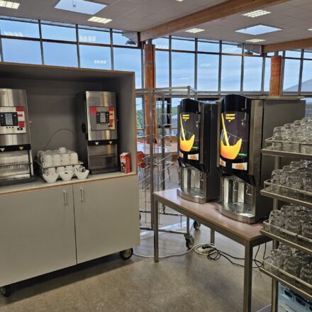 Ein gut beleuchteter Getränkeausgabebereich in einer Kantine mit großen, deckenhohen Fenstern im Hintergrund, durch die viel Tageslicht einfällt. Links stehen zwei silberne Heißgetränkeautomaten mit Tassen, Schälchen und Zubehör wie Zucker bereit. Rechts daneben befinden sich zwei Vending-Maschinen der Marke 