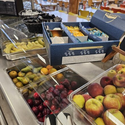 Ein Bereich der Kantine mit einer Auswahl an Getränkezubehör und frischem Obst. Im Vordergrund ist eine geöffnete Teebox mit verschiedenen Sorten Lipton-Tee zu sehen, daneben Körbe mit Zucker- und Süßstoffpäckchen sowie ein Vorrat an Trinkgläsern. Unter der Glasabdeckung liegt eine große Menge Bananen bereit. Im angrenzenden Teil sind Obstbehälter mit frischen Birnen, Orangen, Pflaumen und Äpfeln sichtbar, ordentlich angerichtet in durchsichtigen Kunststoffwannen. Im Hintergrund erkennt man den Speisesaal mit vielen leeren Stühlen und Tischen, was auf eine ruhige Zeit außerhalb der Mahlzeiten hindeutet.