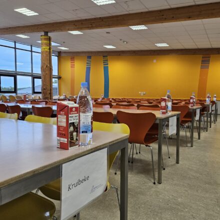 Ein großer Speisesaal mit moderner, schlichter Einrichtung und bunten Akzenten. Die rechte Seite zeigt eine orangefarbene Wand mit farbigen vertikalen Streifen und mehreren langen Tischen mit roten, orangen und gelben Stühlen. Auf den Tischen stehen Wasserflaschen und Milchkartons. Ein Tisch im Vordergrund trägt ein Schild mit der Aufschrift „Kruibeke“. Auf der linken Seite des Bildes sind große Fenster zu sehen, die viel Tageslicht hereinlassen und einen Blick ins Grüne bieten. Holzsäulen und sichtbare Holzbalken tragen zur warmen Atmosphäre des Raums bei. Die Decke ist mit Rasterplatten und eingebauten Leuchten ausgestattet.
