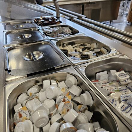 Self-Service-Theke in einer Kantine oder Cafeteria mit verschiedenen Portionsverpackungen. In den Edelstahlschalen befinden sich unter anderem kleine Packungen mit Marmelade, Honig, Butter, Frischkäse (z. B. „Philadelphia“) und Streichkäse („St. Môret“). Im Hintergrund sind weitere Küchenutensilien und große Fenster mit Blick ins Grüne sichtbar.