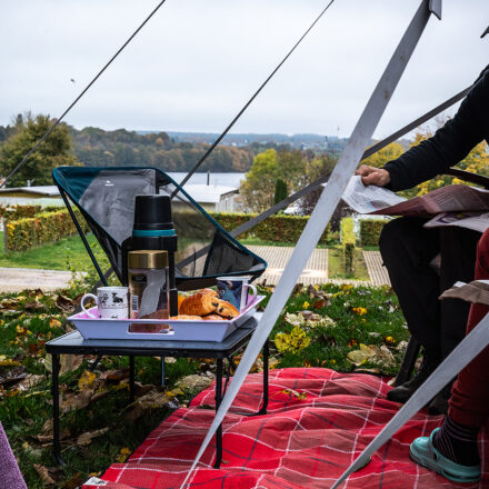 n the foreground is a small folding table with a tray containing a thermos flask, coffee cups and pastries such as croissants. Underneath is a red picnic blanket covered with autumn leaves. A camping chair can be seen in the background on the left, and further back the view opens onto a tranquil landscape with trees and a lake, typical of an autumnal setting. The scenery is relaxed and inviting.