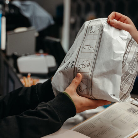 A person hands a customer a paper bag with symbols of biscuits. A menu card and a cash register can be seen in the background, indicating a point of sale such as a restaurant.