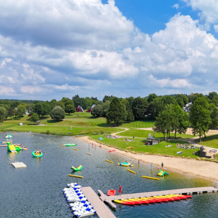 Idyllische Uferlandschaft an einem See, Teil eines Freizeit- oder Ferienparks. Im Vordergrund liegt ein Sandstrand mit mehreren Badenden und Picknickgästen auf der Wiese. Am Steg liegen bunte Kanus und Tretboote (blau-weiß), und im Wasser sind mehrere Spielgeräte und Hindernisse wie ein Wasserpark aufgebaut. Die Umgebung ist sehr grün mit vielen Bäumen und Rasenflächen. Im Hintergrund sieht man mehrere Ferienhäuser oder Chalets mit spitzen Dächern. Rechts im Bild erkennt man ein modernes Gebäude mit Glasfront und eine Spielplatzanlage. Der Himmel ist teils wolkig, aber es scheint ein schöner Sommertag zu sein.