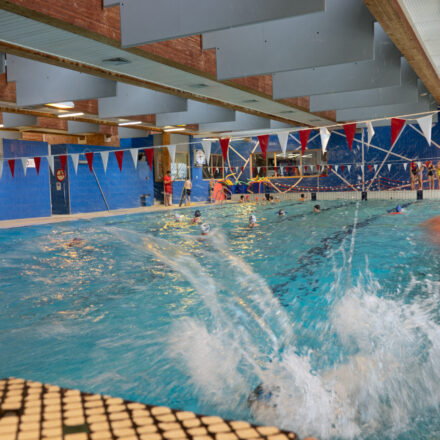 Das Bild zeigt ein lebendiges Schwimmbad-Szenario. Im Vordergrund sieht man spritzendes Wasser, vermutlich durch einen Sprung ins Becken entstanden. Kinder und Jugendliche schwimmen und spielen im Wasser. Im Hintergrund stehen weitere Personen am Beckenrand. Die Decke des Schwimmbads hat eine Konstruktion mit Holz- und Metallelementen. Das Bild vermittelt Spaß, Bewegung und eine sichere Umgebung für Schwimmaktivitäten.