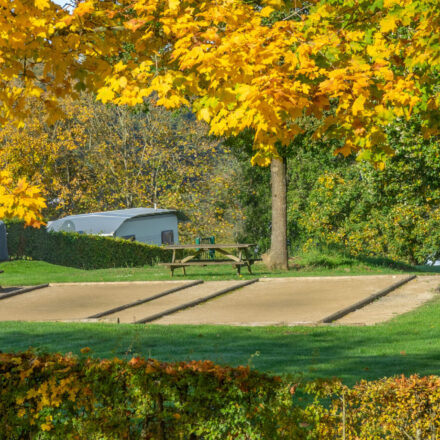 2 Petanquebahnen neben einander in einer herbstlichen Landschaft. Im Hintergrund sieht man das Dach eines Karavans. Es gibt auch einen Picknicktisch und 2 Bänke.