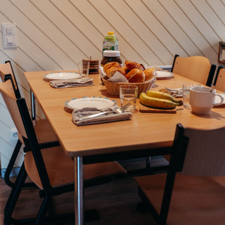 • Frühstücksszene in einem gemütlichen Raum. Auf dem Holztisch stehen mehrere Teller, Besteck, Gläser und Tassen, ergänzt durch einen Korb mit frischen Brötchen. Die Umgebung wirkt schlicht und freundlich, mit Holzverkleidung an den Wänden und einfachen Stühlen, die zu einem Ferienhaus-Ambiente passen. Es entsteht eine familiäre und warme Atmosphäre, die an ein entspanntes Frühstück im Urlaub erinnert.