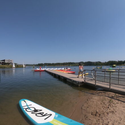 Man sieht im Vordergrund ein Stand-up-Paddling Brett. Es ist auch einen Steg zu sehen, einen See, am ENde des Steges sieht man mehrere Boote: Tretboote und Sit-on-Top Kayaks. Ein Mädchen lauft auf dem Steg. Rechts sieht man mehrere Personen im Wasser auf den Spielgeräten und am Strand.