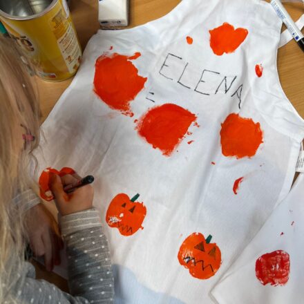 Auf dem Bild sieht man ein Kind, das eine weiße Schürze kreativ gestaltet. Mit orangener Farbe wurden Kürbisse aufgedruckt, die anschließend mit schwarzen und grünen Stiften verziert werden – Gesichter, Stiele und Details. Oben auf der Schürze steht in großen Buchstaben der Name „ELENA“. Auf dem Tisch liegen außerdem Bastelutensilien wie ein Stift, eine Dose und ein Päckchen Taschentücher. Das Bild vermittelt herbstliche Halloween-Stimmung und zeigt eine kreative Bastelaktivität für Kinder.