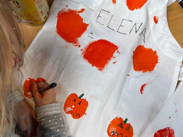 Auf dem Bild sieht man ein Kind, das eine weiße Schürze kreativ gestaltet. Mit orangener Farbe wurden Kürbisse aufgedruckt, die anschließend mit schwarzen und grünen Stiften verziert werden – Gesichter, Stiele und Details. Oben auf der Schürze steht in großen Buchstaben der Name „ELENA“. Auf dem Tisch liegen außerdem Bastelutensilien wie ein Stift, eine Dose und ein Päckchen Taschentücher.
Das Bild vermittelt herbstliche Halloween-Stimmung und zeigt eine kreative Bastelaktivität für Kinder.
