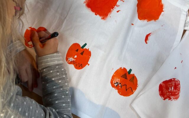 Auf dem Bild sieht man ein Kind, das eine weiße Schürze kreativ gestaltet. Mit orangener Farbe wurden Kürbisse aufgedruckt, die anschließend mit schwarzen und grünen Stiften verziert werden – Gesichter, Stiele und Details. Oben auf der Schürze steht in großen Buchstaben der Name „ELENA“. Auf dem Tisch liegen außerdem Bastelutensilien wie ein Stift, eine Dose und ein Päckchen Taschentücher.
Das Bild vermittelt herbstliche Halloween-Stimmung und zeigt eine kreative Bastelaktivität für Kinder.