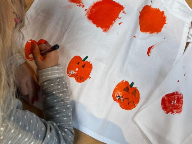 Auf dem Bild sieht man ein Kind, das eine weiße Schürze kreativ gestaltet. Mit orangener Farbe wurden Kürbisse aufgedruckt, die anschließend mit schwarzen und grünen Stiften verziert werden – Gesichter, Stiele und Details. Oben auf der Schürze steht in großen Buchstaben der Name „ELENA“. Auf dem Tisch liegen außerdem Bastelutensilien wie ein Stift, eine Dose und ein Päckchen Taschentücher.
Das Bild vermittelt herbstliche Halloween-Stimmung und zeigt eine kreative Bastelaktivität für Kinder.