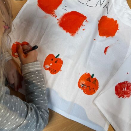 Auf dem Bild sieht man ein Kind, das eine weiße Schürze kreativ gestaltet. Mit orangener Farbe wurden Kürbisse aufgedruckt, die anschließend mit schwarzen und grünen Stiften verziert werden – Gesichter, Stiele und Details. Oben auf der Schürze steht in großen Buchstaben der Name „ELENA“. Auf dem Tisch liegen außerdem Bastelutensilien wie ein Stift, eine Dose und ein Päckchen Taschentücher. Das Bild vermittelt herbstliche Halloween-Stimmung und zeigt eine kreative Bastelaktivität für Kinder.