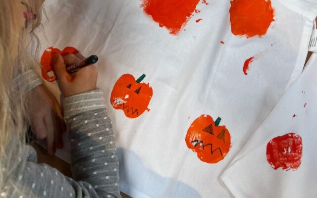 Auf dem Bild sieht man ein Kind, das eine weiße Schürze kreativ gestaltet. Mit orangener Farbe wurden Kürbisse aufgedruckt, die anschließend mit schwarzen und grünen Stiften verziert werden – Gesichter, Stiele und Details. Oben auf der Schürze steht in großen Buchstaben der Name „ELENA“. Auf dem Tisch liegen außerdem Bastelutensilien wie ein Stift, eine Dose und ein Päckchen Taschentücher. Das Bild vermittelt herbstliche Halloween-Stimmung und zeigt eine kreative Bastelaktivität für Kinder.