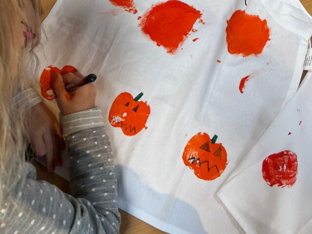 Auf dem Bild sieht man ein Kind, das eine weiße Schürze kreativ gestaltet. Mit orangener Farbe wurden Kürbisse aufgedruckt, die anschließend mit schwarzen und grünen Stiften verziert werden – Gesichter, Stiele und Details. Oben auf der Schürze steht in großen Buchstaben der Name „ELENA“. Auf dem Tisch liegen außerdem Bastelutensilien wie ein Stift, eine Dose und ein Päckchen Taschentücher.
Das Bild vermittelt herbstliche Halloween-Stimmung und zeigt eine kreative Bastelaktivität für Kinder.