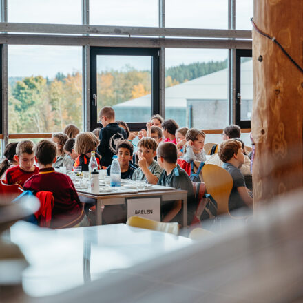 Eine Gruppe von Kindern sitzt in einer hellen, modernen Mensa an einem langen Tisch beim gemeinsamen Essen. Auf dem Tisch stehen leere Teller, Besteck und Wasserflaschen. Am Tisch hängt ein Schild mit der Aufschrift „BAELEN“, vermutlich der Name der Gruppe oder Schule. Im Hintergrund sind große Fenster mit Blick auf herbstlich gefärbte Bäume und ein Gebäude zu sehen. Die Stimmung wirkt lebendig und gesellig.
