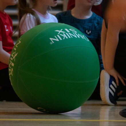 Ein großer grüner Ball mit der Aufschrift ‚OMNIKIN SIX‘ liegt auf dem Boden einer Sporthalle. Um den Ball herum sitzen mehrere Kinder in Sportkleidung.
