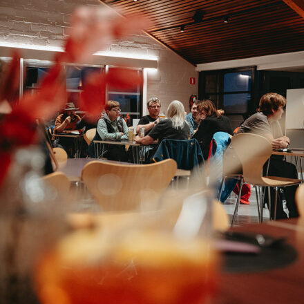 Blick auf eine Gruppe von Menschen, die sich gemütlich unterhalten. Die Elemente im Vordergrund sind verschwommen (eine Pflanze und ein Glas mit einem Aperitif).