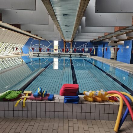 Das Foto zeigt das 25-Meter-Hallenbad in Worriken. Im Vordergrund liegen bunte Schwimmhilfen ordentlich aufgereiht am Beckenrand – darunter Schwimmbretter, Poolnudeln, Schwimmflügel und andere Trainingsutensilien. Das Wasser im Becken ist klar und ruhig, die schwarzen Markierungslinien für das Bahnschwimmen sind gut sichtbar. Über dem Becken hängen Wimpelketten zur Orientierung. Die Architektur ist modern und funktional: Die linke Seite besteht aus großen Fensterflächen, durch die viel Tageslicht ins Bad fällt. Die Wände sind in einem kräftigen Blau gehalten, was dem Raum eine sportliche Atmosphäre verleiht.