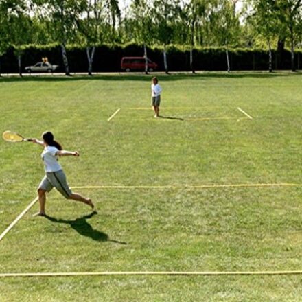 Zwei Personen spielen Badminton ohne Netz auf einer großen Rasenfläche im Freien. Die Spielfelder sind mit gelbem Band auf dem Gras markiert. Eine Person im Vordergrund schlägt den Federball, während die andere Person im Hintergrund bereitsteht. Im Hintergrund sind Bäume, ein Zaun und parkende Autos zu sehen.