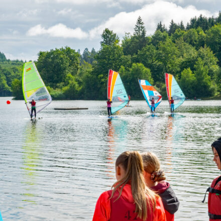 Das Bild zeigt eine Gruppe von Kindern und Jugendlichen, die beim Windsurfen auf einem See aktiv sind. Im Vordergrund stehen einige Teilnehmer mit Schwimmwesten am Ufer und beobachten die Surfer auf dem Wasser. Im Hintergrund gleiten mehrere Windsurfer mit bunten Segeln – in Grün, Orange und Blau – über die Wasseroberfläche. Die Szenerie ist von dichtem Wald und grüner Natur umgeben, während der Himmel leicht bewölkt ist, aber auch Sonnenstrahlen durchscheinen. Es wirkt wie ein aktiver, fröhlicher Sommertag, vermutlich während eines Sport- oder Ferienlagers.