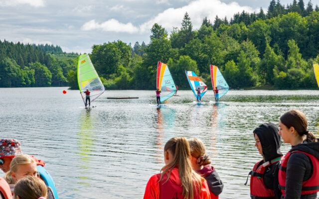 Das Bild zeigt eine Gruppe von Kindern und Jugendlichen, die beim Windsurfen auf einem See aktiv sind. Im Vordergrund stehen einige Teilnehmer mit Schwimmwesten am Ufer und beobachten die Surfer auf dem Wasser. Im Hintergrund gleiten mehrere Windsurfer mit bunten Segeln – in Grün, Orange und Blau – über die Wasseroberfläche. Die Szenerie ist von dichtem Wald und grüner Natur umgeben, während der Himmel leicht bewölkt ist, aber auch Sonnenstrahlen durchscheinen. Es wirkt wie ein aktiver, fröhlicher Sommertag, vermutlich während eines Sport- oder Ferienlagers.