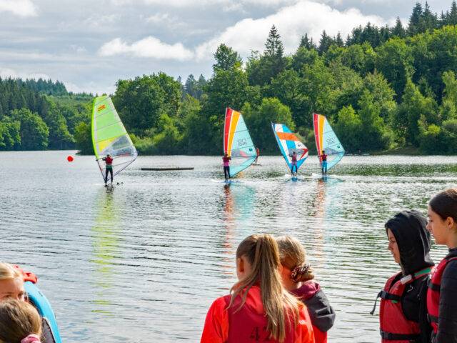 Das Bild zeigt eine Gruppe von Kindern und Jugendlichen, die beim Windsurfen auf einem See aktiv sind. Im Vordergrund stehen einige Teilnehmer mit Schwimmwesten am Ufer und beobachten die Surfer auf dem Wasser. Im Hintergrund gleiten mehrere Windsurfer mit bunten Segeln – in Grün, Orange und Blau – über die Wasseroberfläche. Die Szenerie ist von dichtem Wald und grüner Natur umgeben, während der Himmel leicht bewölkt ist, aber auch Sonnenstrahlen durchscheinen. Es wirkt wie ein aktiver, fröhlicher Sommertag, vermutlich während eines Sport- oder Ferienlagers.