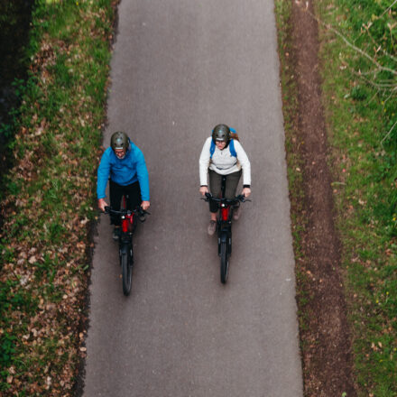 Das Bild zeigt zwei Personen, die nebeneinander auf einem asphaltierten Radweg durch eine grüne, naturbelassene Umgebung fahren. Beide tragen Fahrradhelme – die linke Person eine blaue Jacke, die rechte eine weiße Jacke und einen blauen Rucksack. Sie fahren auf modernen Fahrrädern oder E-Bikes mit roten Elementen. Das Bild wurde von oben aufgenommen und vermittelt eine ruhige, entspannte Stimmung – ideal für eine Fahrradtour in der Natur. Links und rechts des Weges sind Gras, Laub und Waldanteile zu sehen.