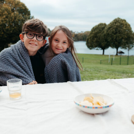 2 Kinder mit Decke. Im Vordergrund gibt es einen Tisch im Hintergrund einen See. Die Kinder lachen. Es sieht aus wie eine Aperitifszene im Freiem mit einem Weinglas, Chs und Glas mit orange Limo.