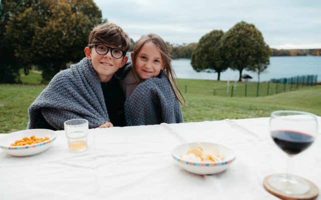 2 Kinder mit Decke. Im Vordergrund gibt es einen Tisch im Hintergrund einen See. Die Kinder lachen. Es sieht aus wie eine Aperitifszene im Freiem mit einem Weinglas, Chs und Glas mit orange Limo.