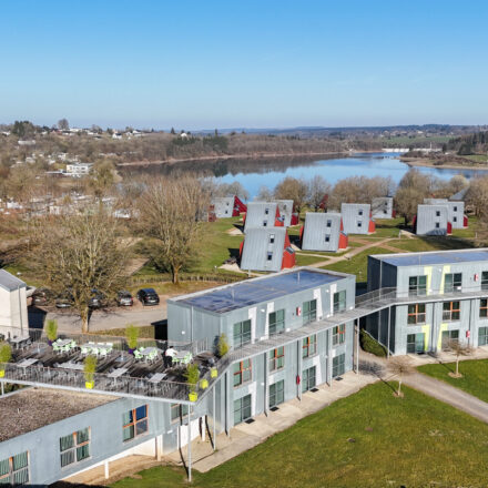 Vue aérienne de Worriken, un centre de sport et de vacances en Belgique. Au premier plan, on voit des bâtiments modernes, dont le B&B Sporthouse et sa terrasse extérieure. Derrière se trouvent les maisons de vacances caractéristiques (chalets) au milieu d'un paysage verdoyant. En arrière-plan s'étend le lac idyllique de Bütgenbach, entouré d'arbres et du village de Bütgenbach. Sur la gauche de l'image, on reconnaît en outre une partie de la zone de camping. Le temps ensoleillé et le ciel bleu confèrent à la scène une atmosphère accueillante.